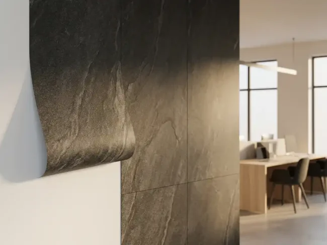 Steinoptik-Folie an Bürowand mit angehobenem Rand, der weiße Wand darunter freilegt, anthrazitfarbene Naturssteintöne, minimalistisches Interieur im Hintergrund.